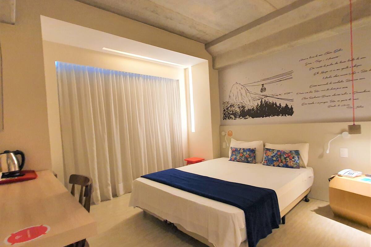 A hotel room with a bed, a desk, and a chair. There is a window with curtains, a wall with a drawing, and a wall-mounted lamp.