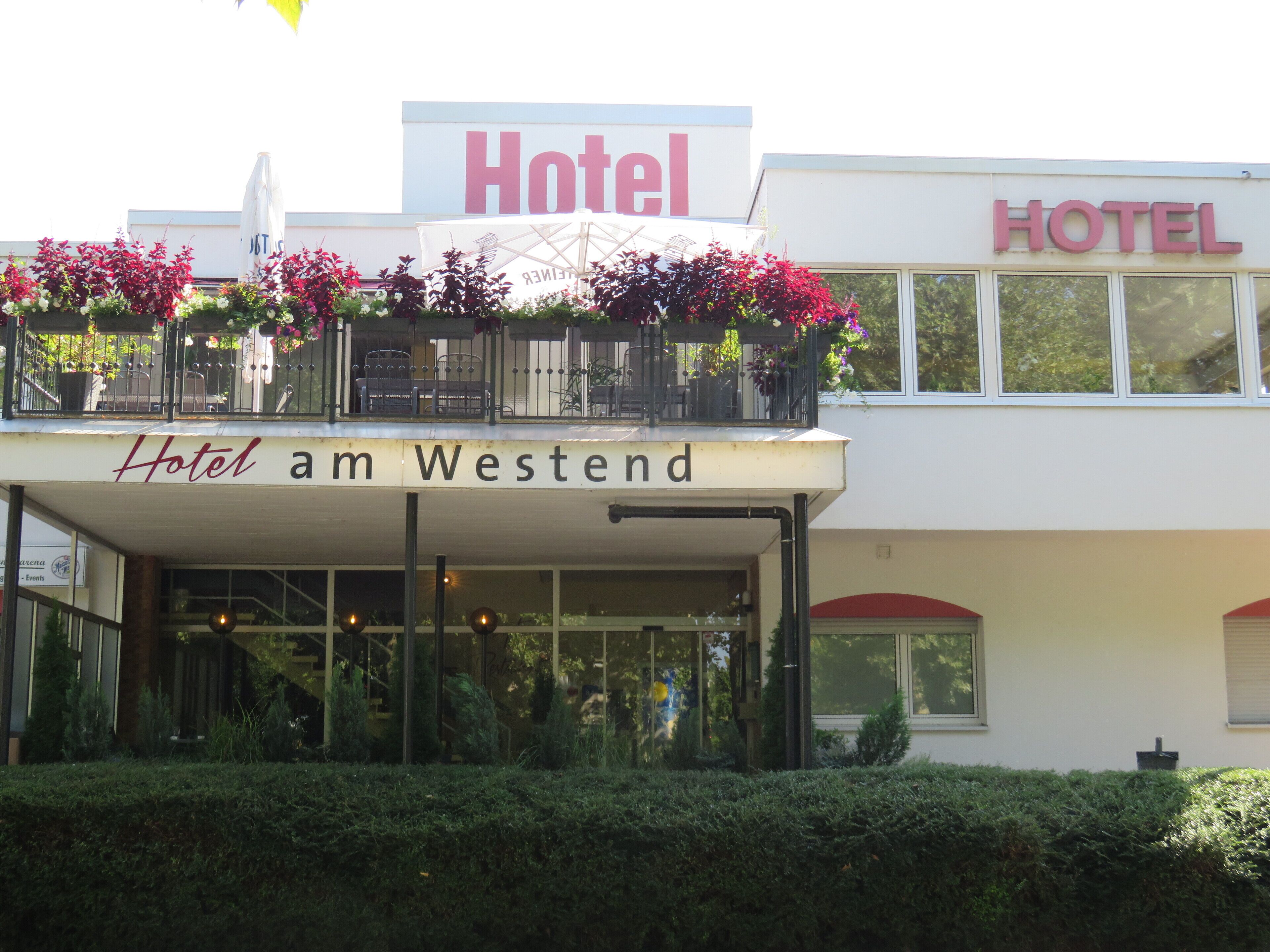 A hotel with a balcony adorned with flowers, a sign reading "Hotel am Westend," and a clear view of the entrance.