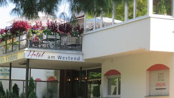 Hotelli, jonka parvekkeella on runsaasti kukkia, ja jonka yläpuolella on kyltti, jossa lukee ”Hotel am Westend”, sekä selkeästi näkyvä sisäänkäynti, jonka yläpuolella on kyltti, jossa lukee ”Hotel Eingang”.