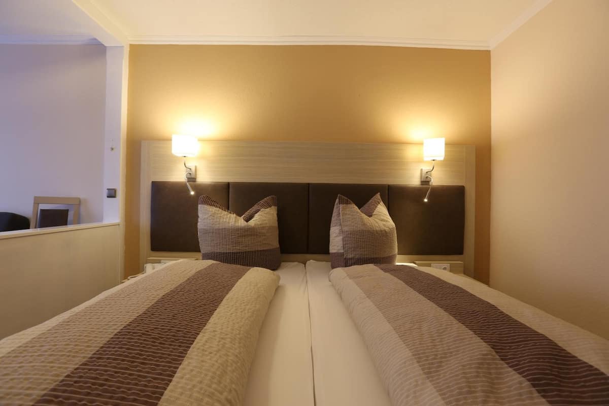 Quarto de hotel com duas camas, cabeceira com luminárias fixadas na parede e espelho sobre as camas.