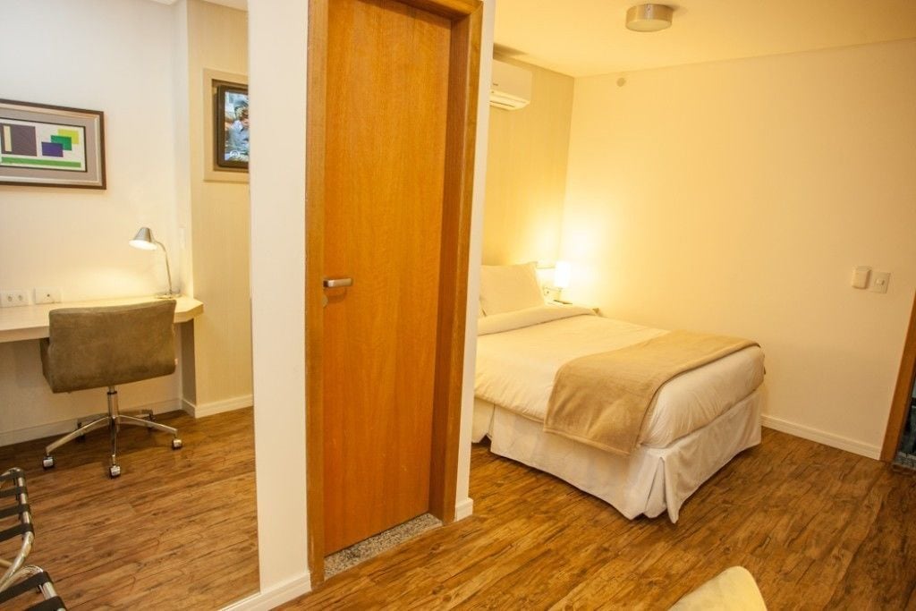 Un dormitorio con suelo de madera, una cama con ropa de cama blanca, un escritorio con una silla y una puerta.
