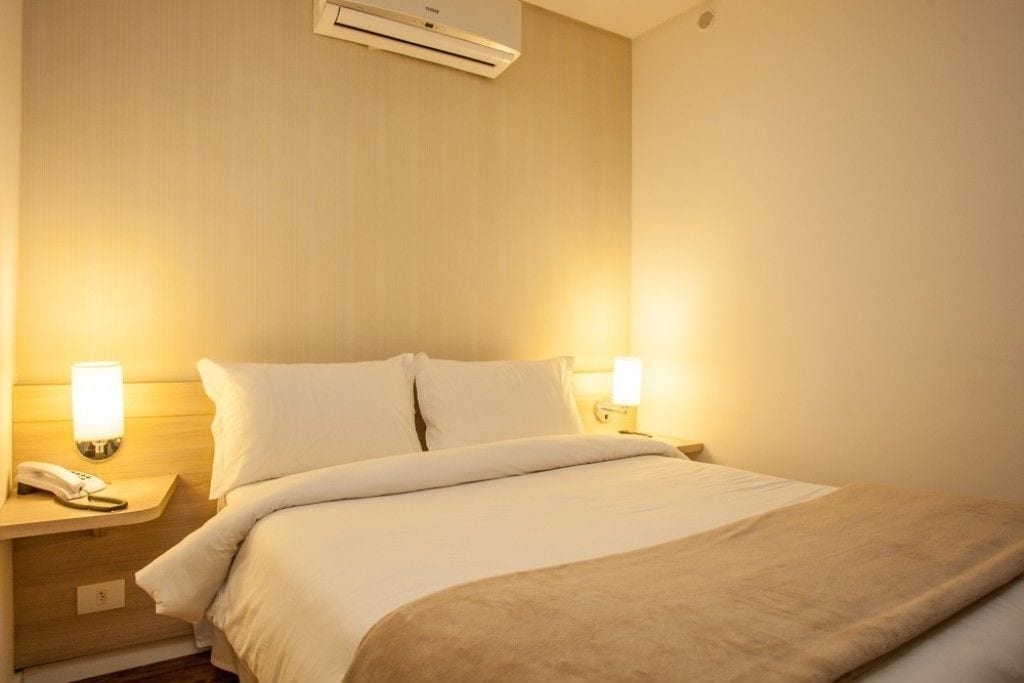 A hotel room with a bed, bedside lamps, a phone, and an air conditioning unit.