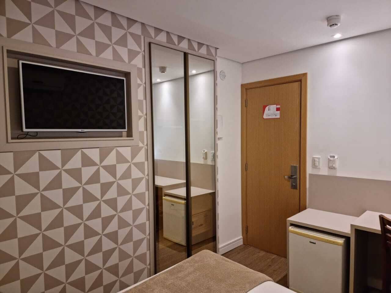 Une chambre d’hôtel de taille réduite avec un mur orné d’un motif géométrique, une porte coulissante donnant accès à la salle de bain, un petit bureau et un mini-réfrigérateur.
