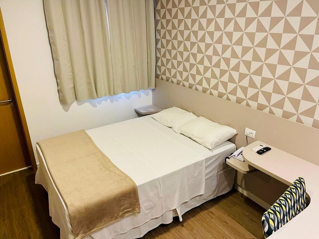 Una cama bien hecha con una manta color beige, una mesita de noche con un teléfono y un papel tapiz estampado.