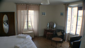 A bedroom with a large bed, a chair, a small table, a dresser, and a window with curtains. - Le Voltaire Chambres et Table d'Hotes (Airvault)
