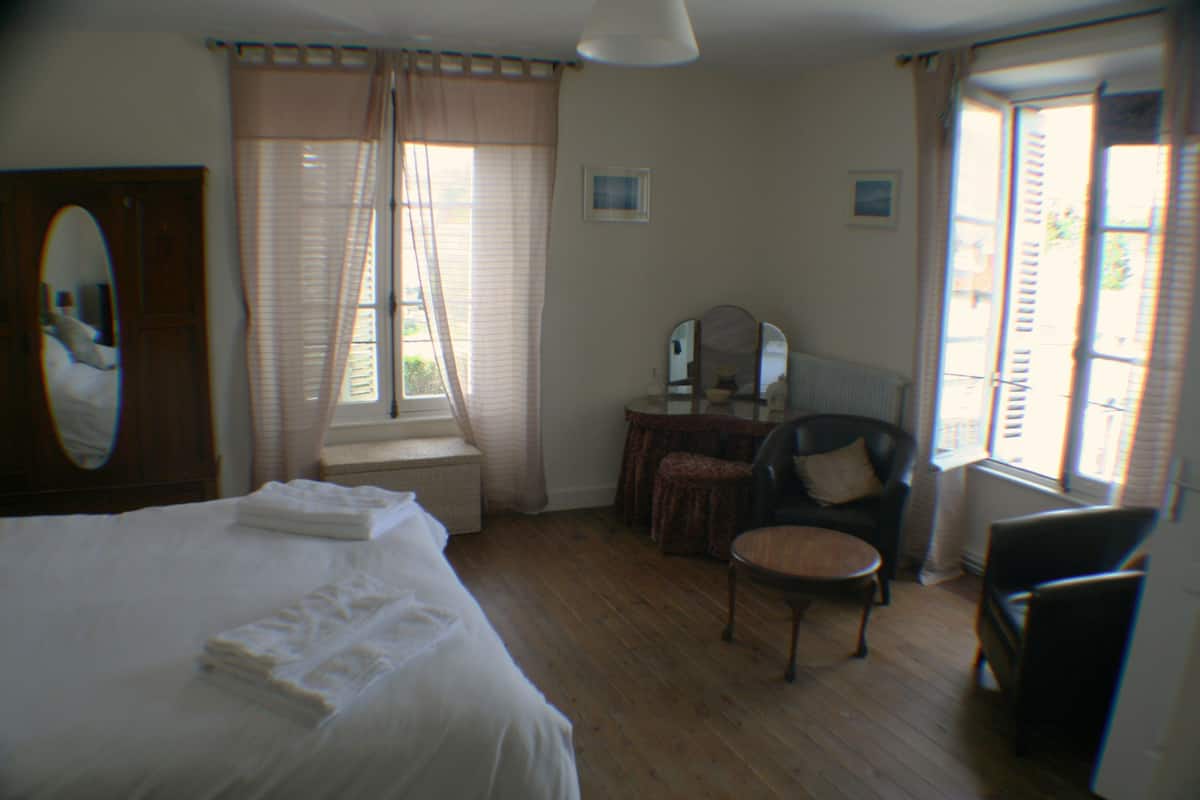 Quarto com uma cama grande, uma cadeira, uma mesinha, uma cômoda e uma janela com cortinas.