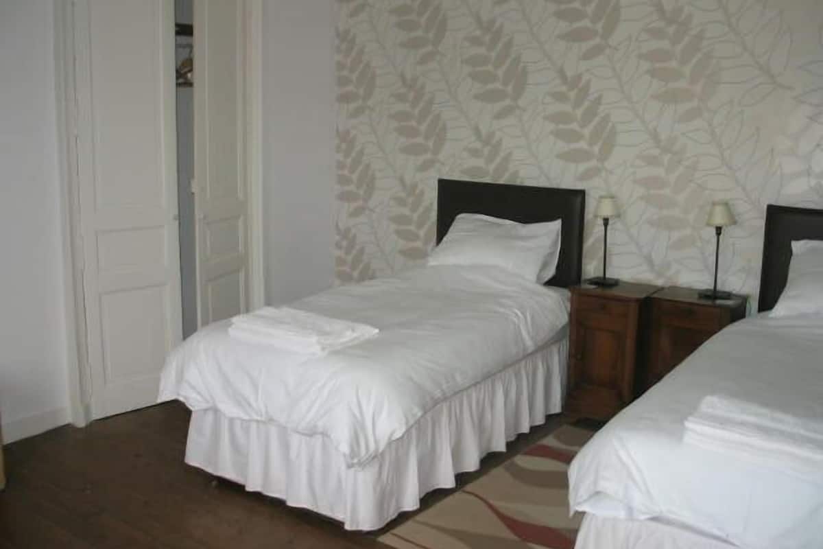 Cama de casal com lençóis brancos, mesa de cabeceira com abajur e papel de parede estampado.