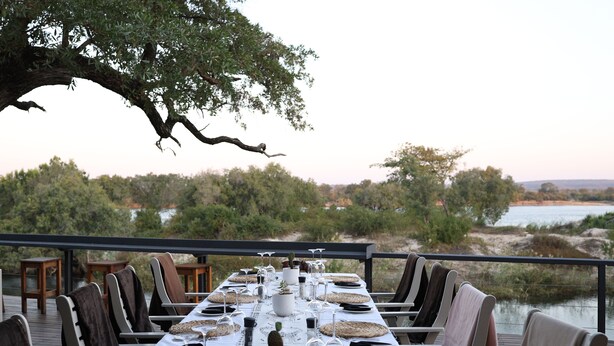 An outdoor dining area with a long table set for a meal, overlooking a natural landscape with trees and a body of water.