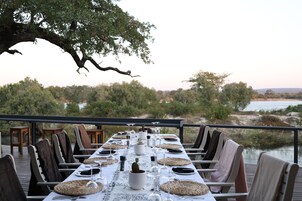 Un área de comedor al aire libre con una mesa larga dispuesta para una comida, con vistas a un paisaje natural con árboles y una masa de agua.