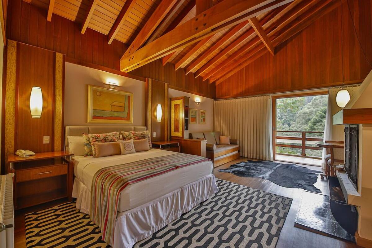 Une chambre spacieuse avec un grand lit, des poutres apparentes en bois au plafond, une grande fenêtre offrant une belle vue et un bureau avec une lampe.