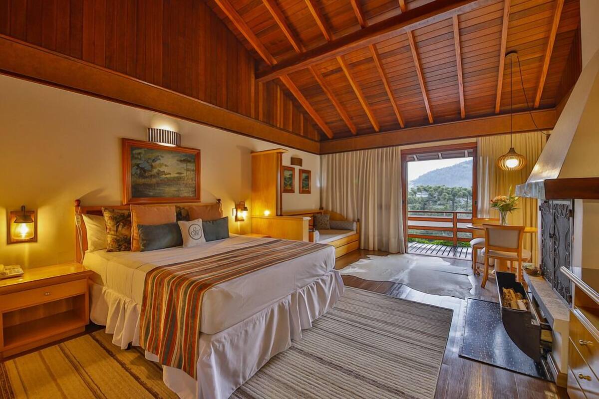 Une chambre spacieuse avec un grand lit, des poutres apparentes en bois au plafond, un balcon avec vue et une cheminée.