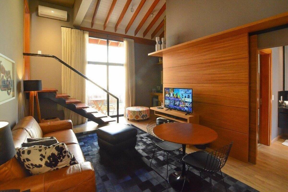 Un salon moderne avec un canapé, un pouf et une table basse. On y trouve un téléviseur posé sur un meuble en bois, ainsi qu’un escalier menant à un autre niveau.