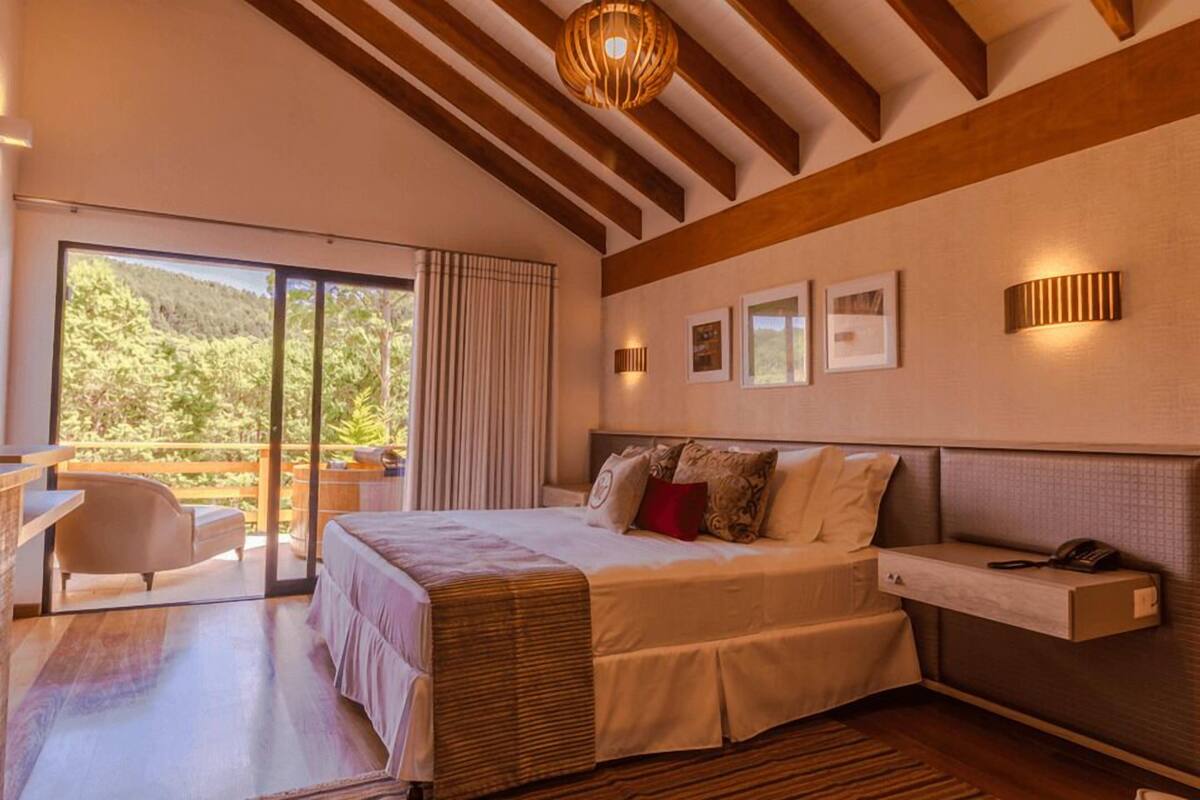 Une chambre dotée d’un grand lit, d’un bureau et d’une chaise, d’un balcon offrant une vue sur les arbres et de poutres en bois au plafond.