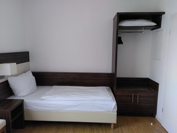 Una cama individual con cabecera de madera y una mesita de noche pequeña. A la derecha de la cama, un armario con espejo y barra para colgar ropa.