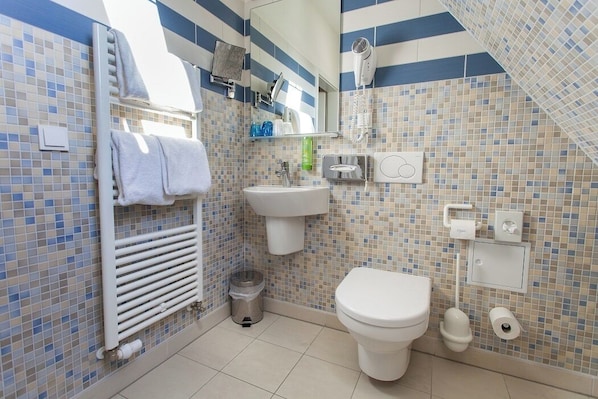 Un baño con un mosaico de azulejos en la pared sobre el lavamanos, un lavamanos blanco, un inodoro, un perchero para toallas, un secador de pelo y un cubo de basura.