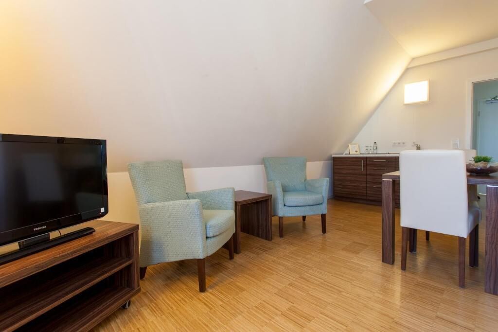 Una sala de estar con una televisión de pantalla plana sobre un soporte de madera, dos sillones azul claro, una mesita y un comedor con una silla blanca y una mesa de madera.