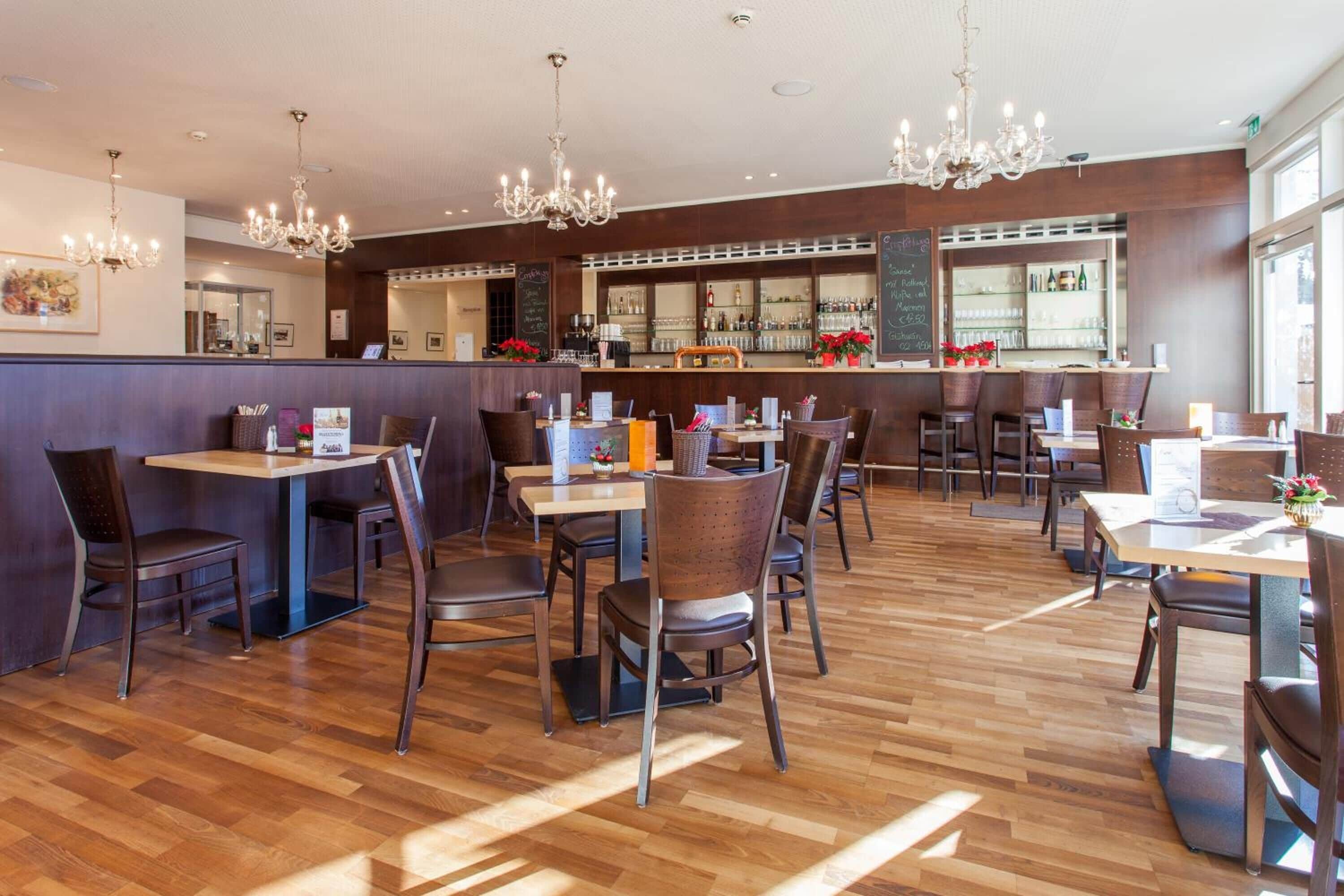 A modern restaurant with wooden flooring, multiple tables and chairs, and a bar area with shelves of bottles.