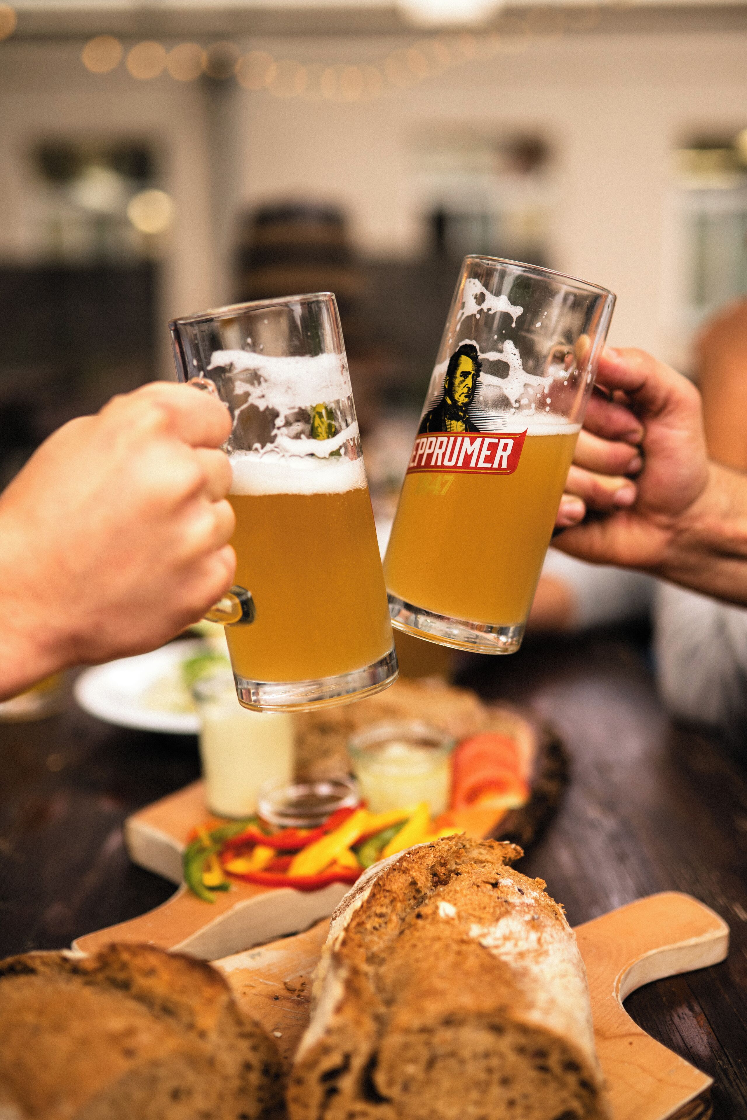 Dos personas sosteniendo vasos de cerveza con espuma, un pan y una tabla de cortar con verduras sobre una mesa de madera.