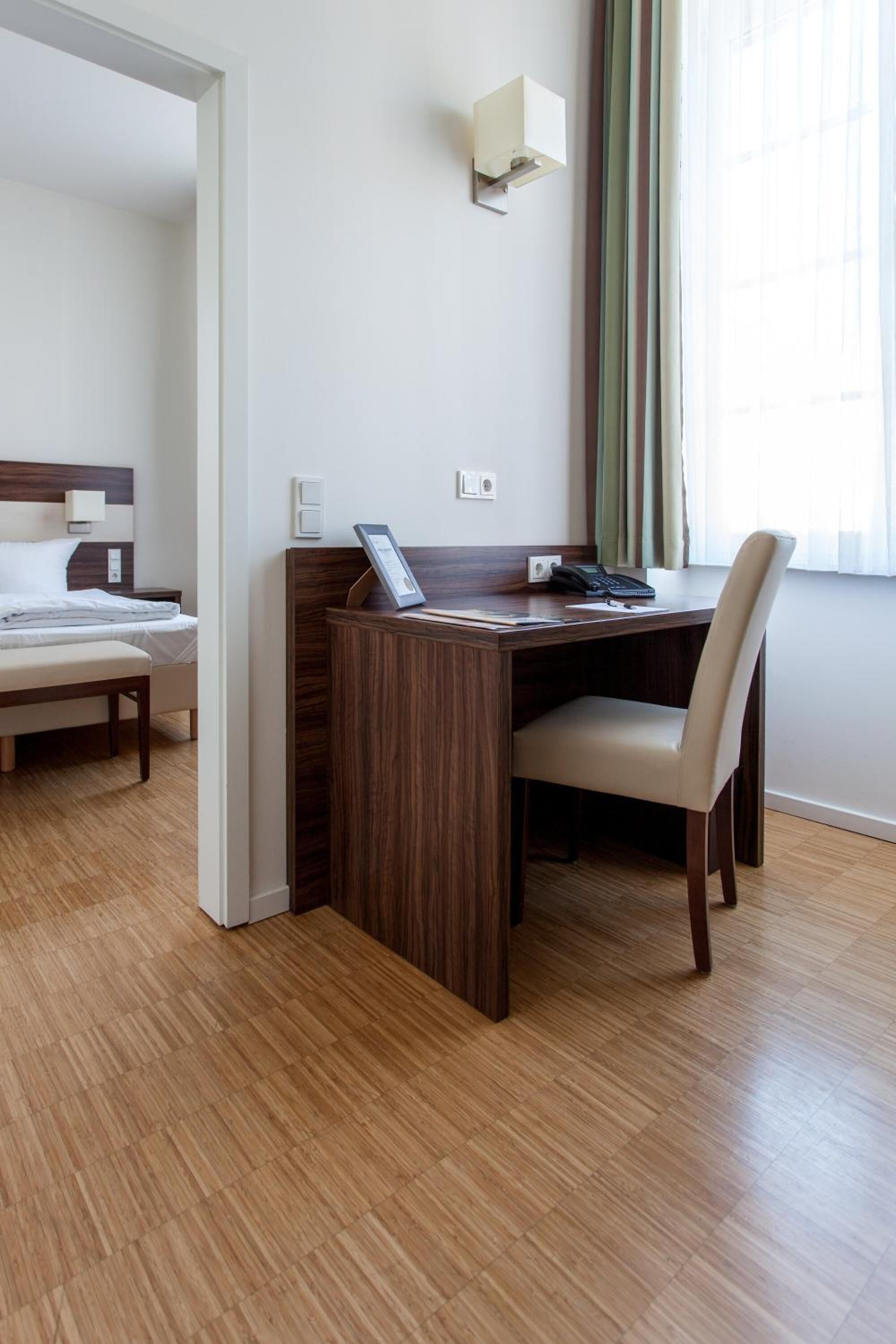 Una habitación de hotel con un escritorio de madera, una silla, una cama y un ventanal con cortinas.