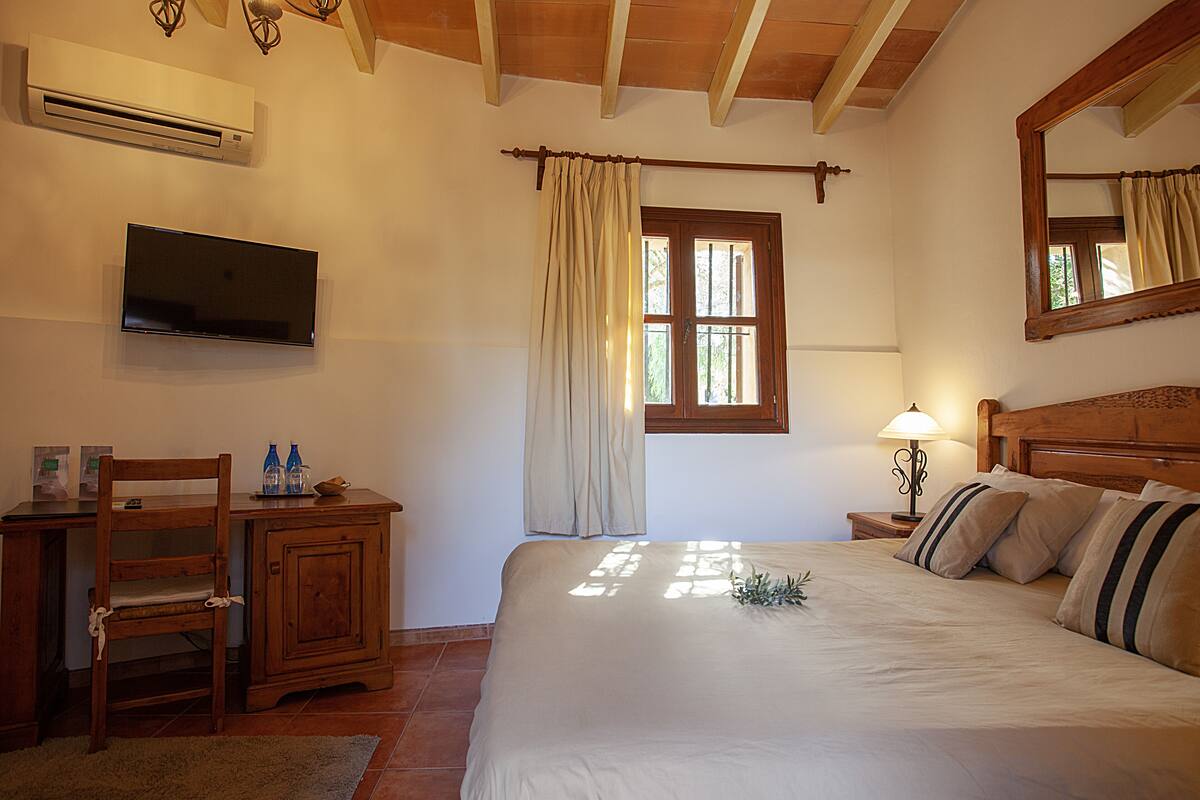 Une chambre à coucher comprenant un lit, un bureau avec une chaise, un téléviseur fixé au mur, une fenêtre avec des rideaux et une table de chevet avec une lampe.