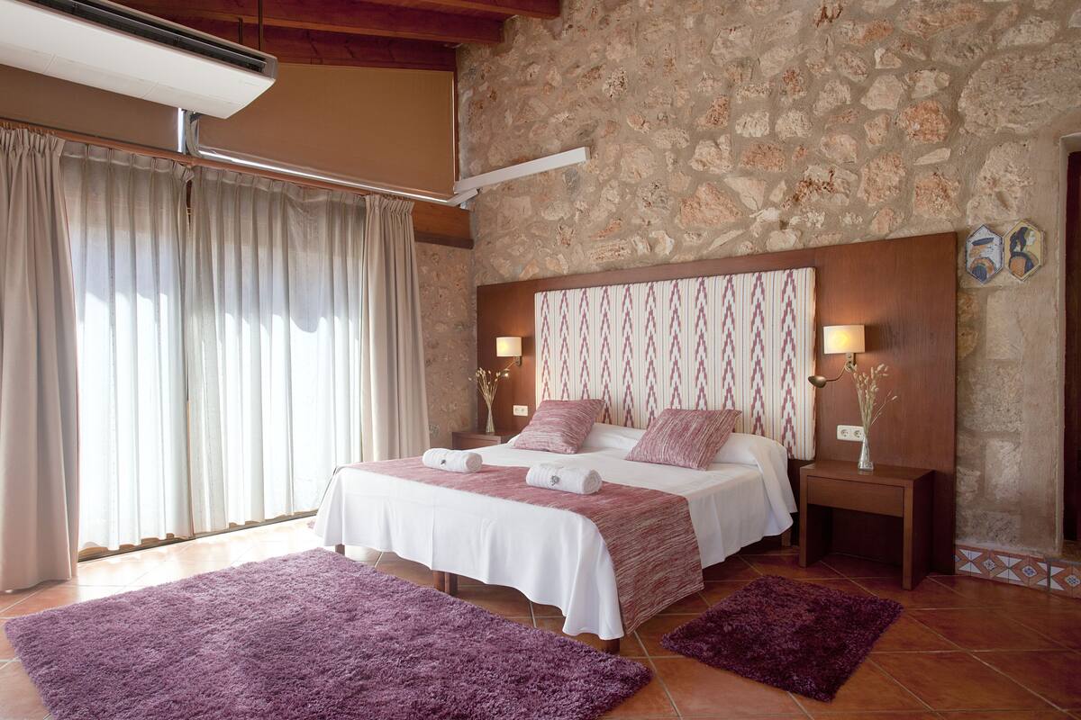Une chambre avec un mur en pierre, un grand lit avec une literie blanche et rose, deux tables de chevet avec des lampes et un tapis violet.