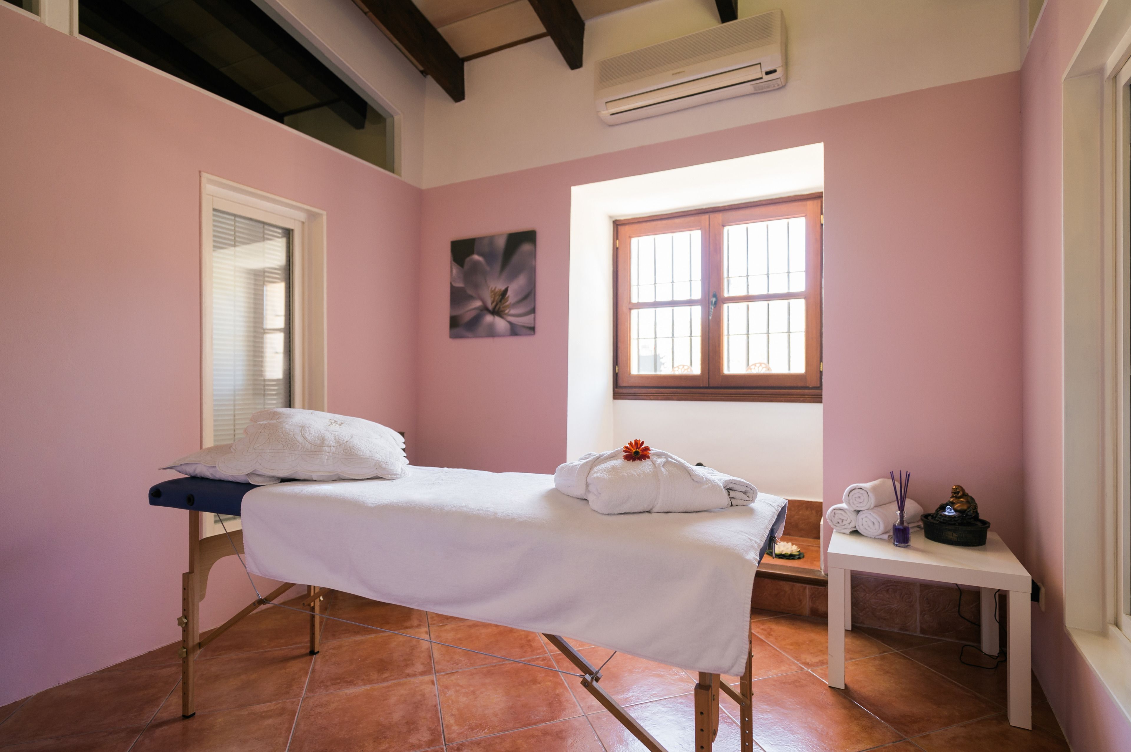 A room with a bed, a window, a painting, and a small table with a statue and incense sticks.