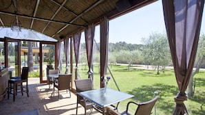 A covered patio with wicker furniture, a glass table, and a view of a grassy landscape.