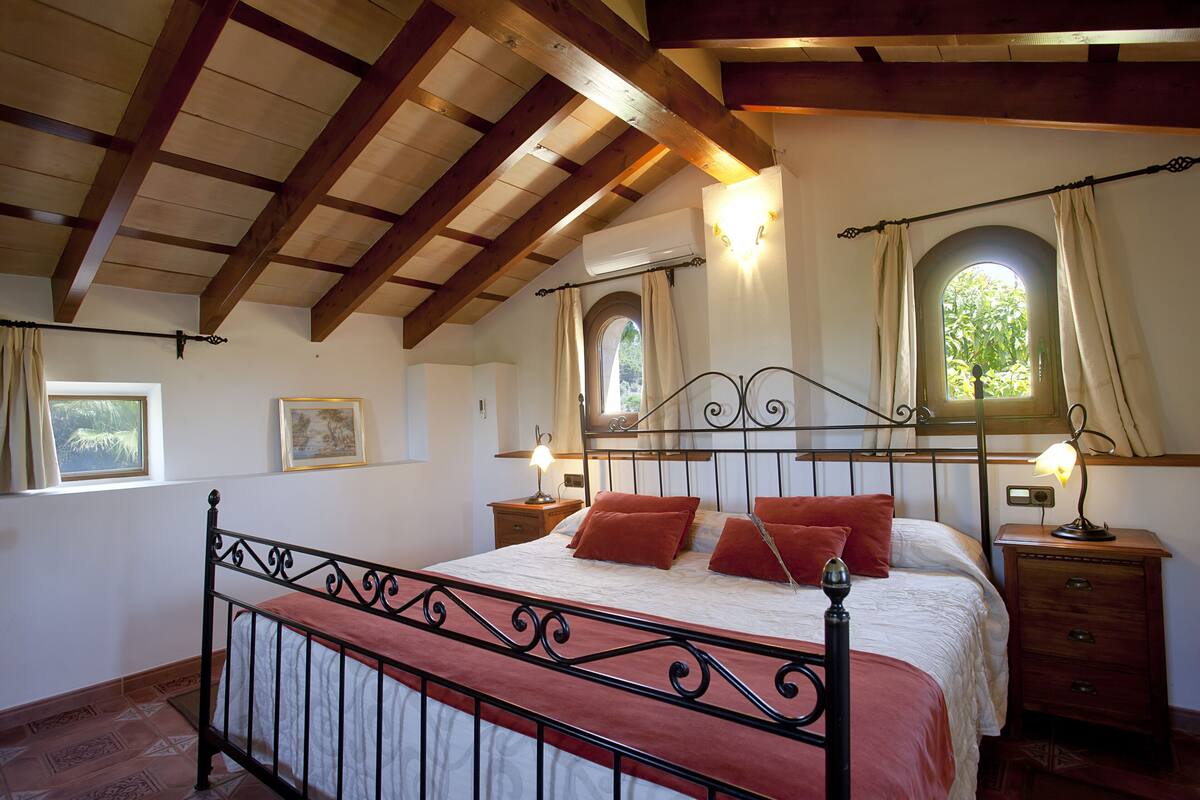 Une chambre avec un plafond en bois, un lit avec un cadre en métal, deux tables de chevet avec des lampes et une fenêtre avec des rideaux.