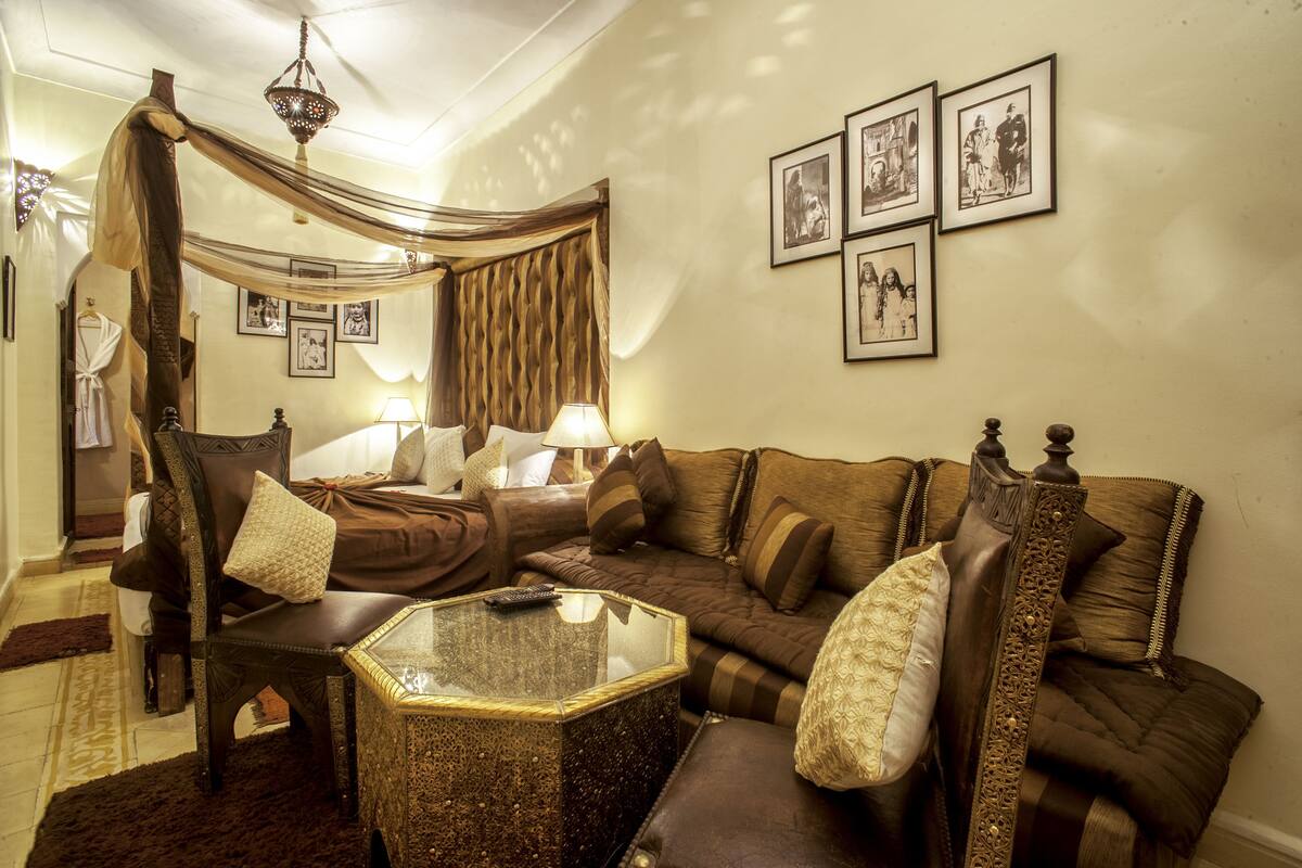 A cozy room with a four-poster bed, a large sofa, and a glass coffee table. There are framed pictures on the wall and a chandelier hanging from the ceiling.