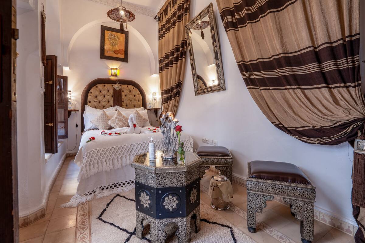 A bedroom with a large bed, a nightstand, a small table, and a bench.