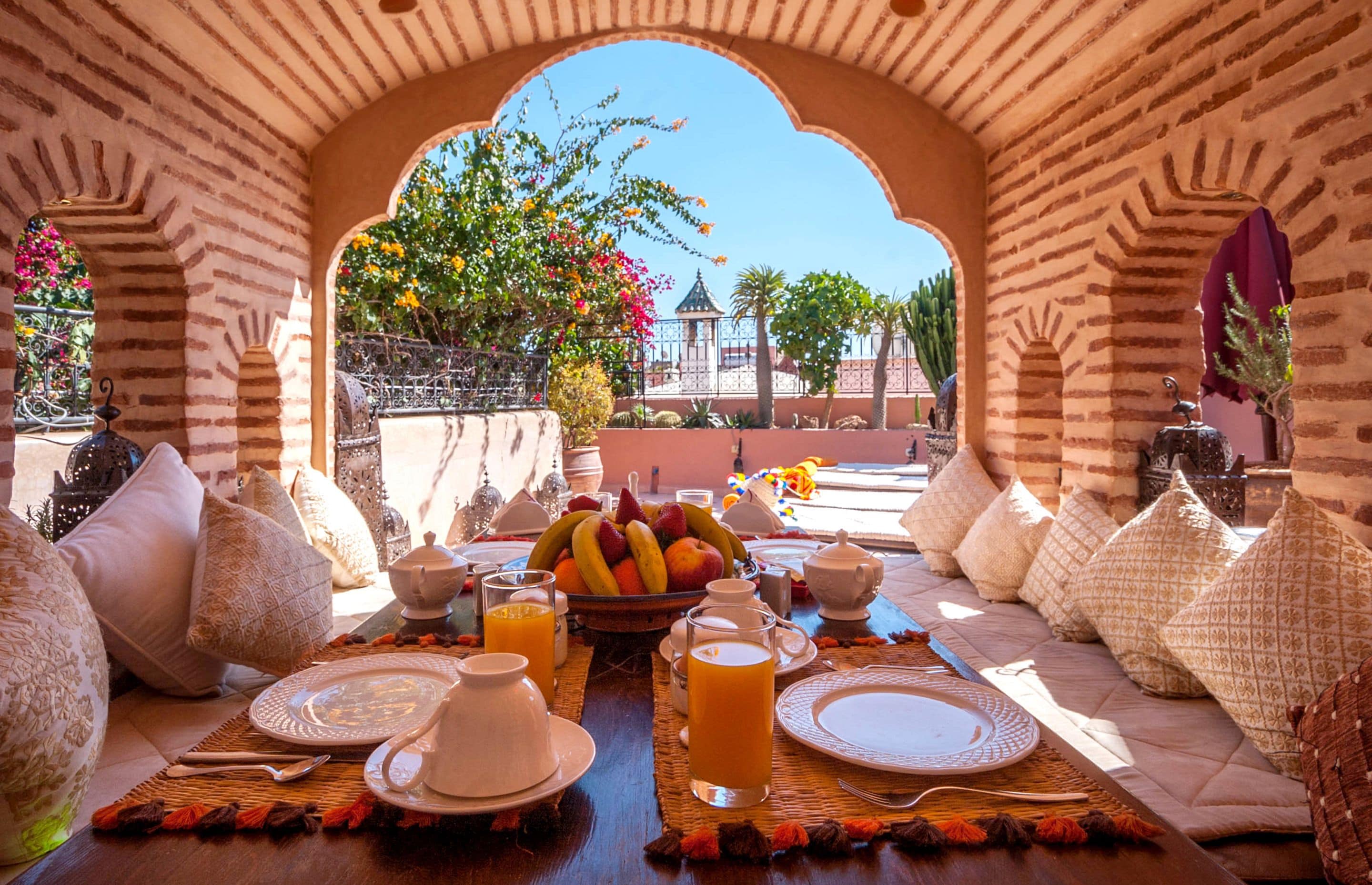 A breakfast setup with a fruit bowl, tea set, and orange juice on a table under a brick archway.