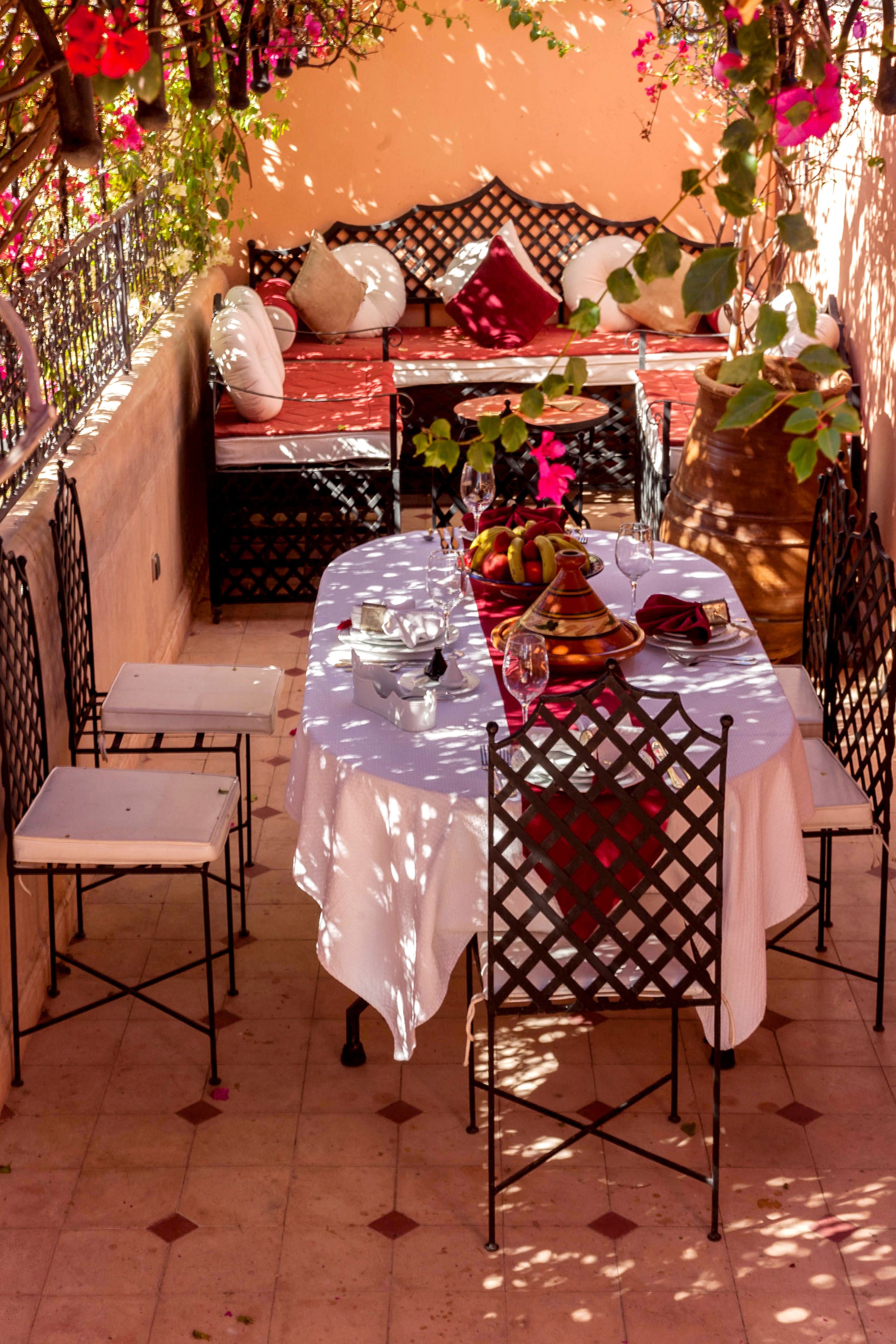 Un área de comedor al aire libre con una mesa preparada para dos, rodeada de sillas, un banco acolchado y una pared decorativa con plantas trepadoras.