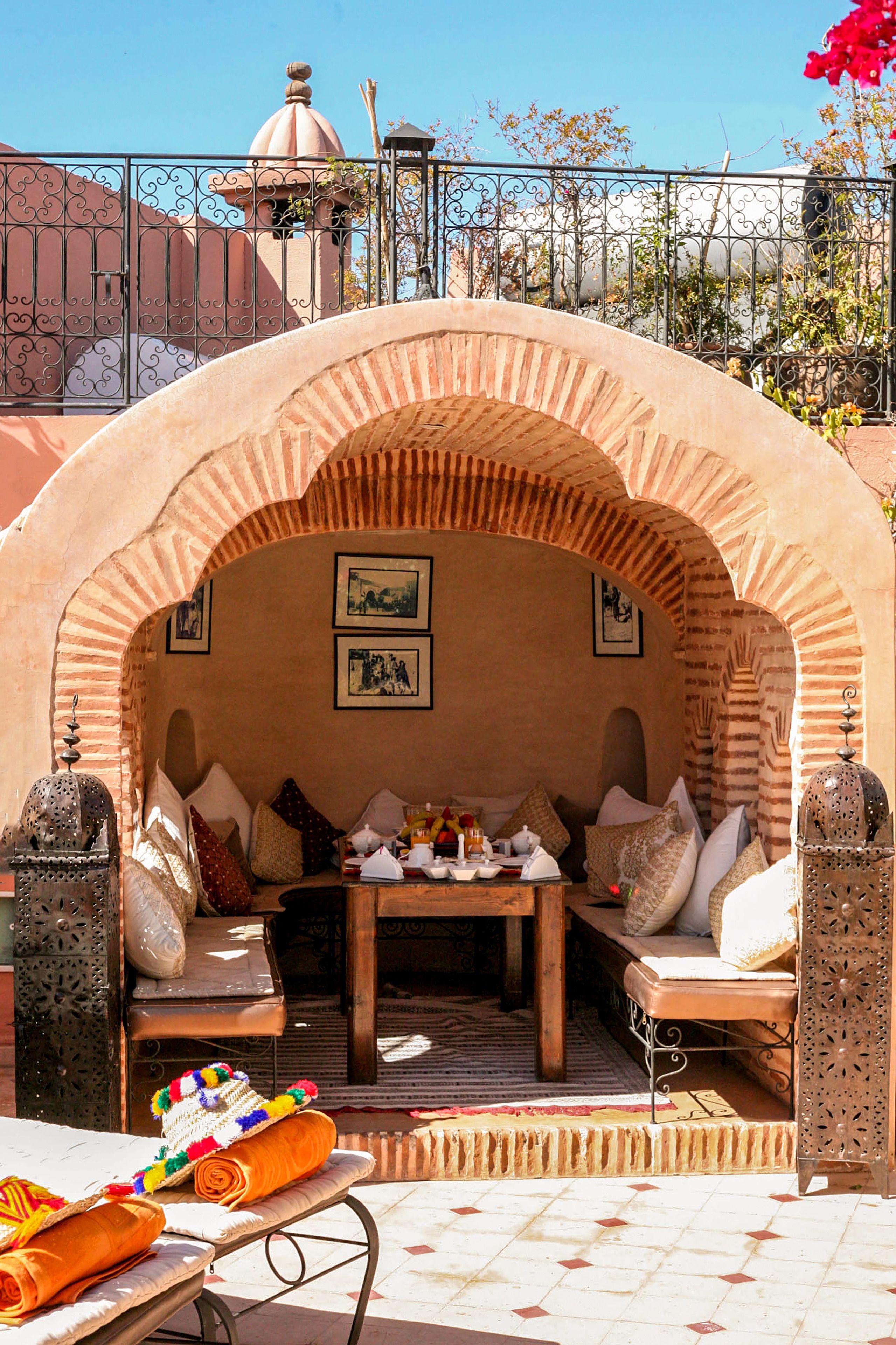 An outdoor seating area with a stone arch, cushioned benches, and a table set with various items.