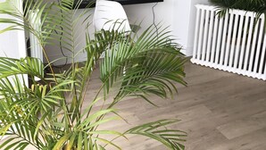Una habitación con tres estantes negros contra una pared blanca, cada uno con libros, un reloj y una vela. Frente a los estantes hay una silla blanca y, a la derecha, una valla blanca con plantas.