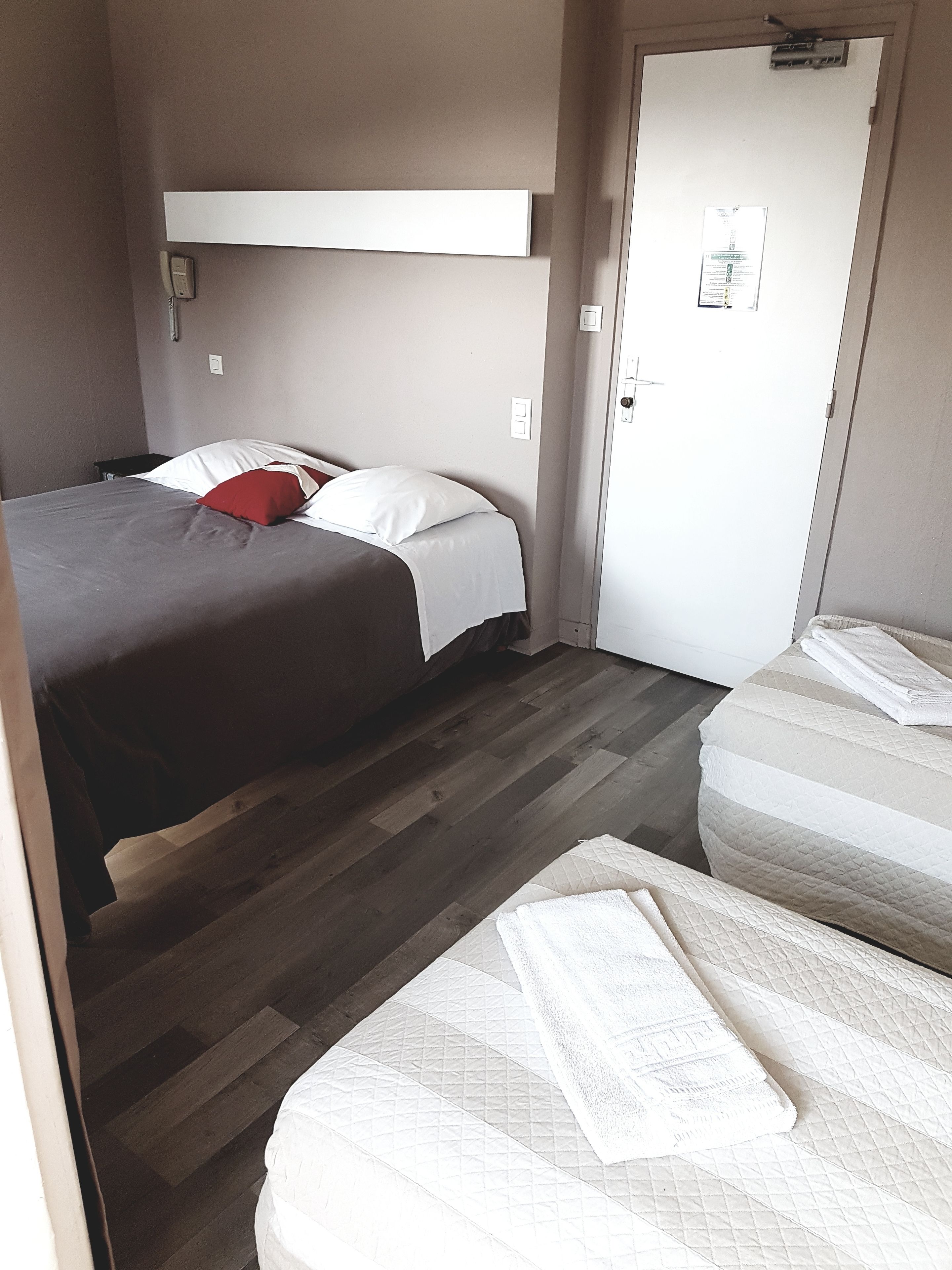 Habitación de hotel con dos camas, una con almohada roja y la otra con ropa blanca. Se observa una puerta con cerradura y un interruptor de luz en la pared.