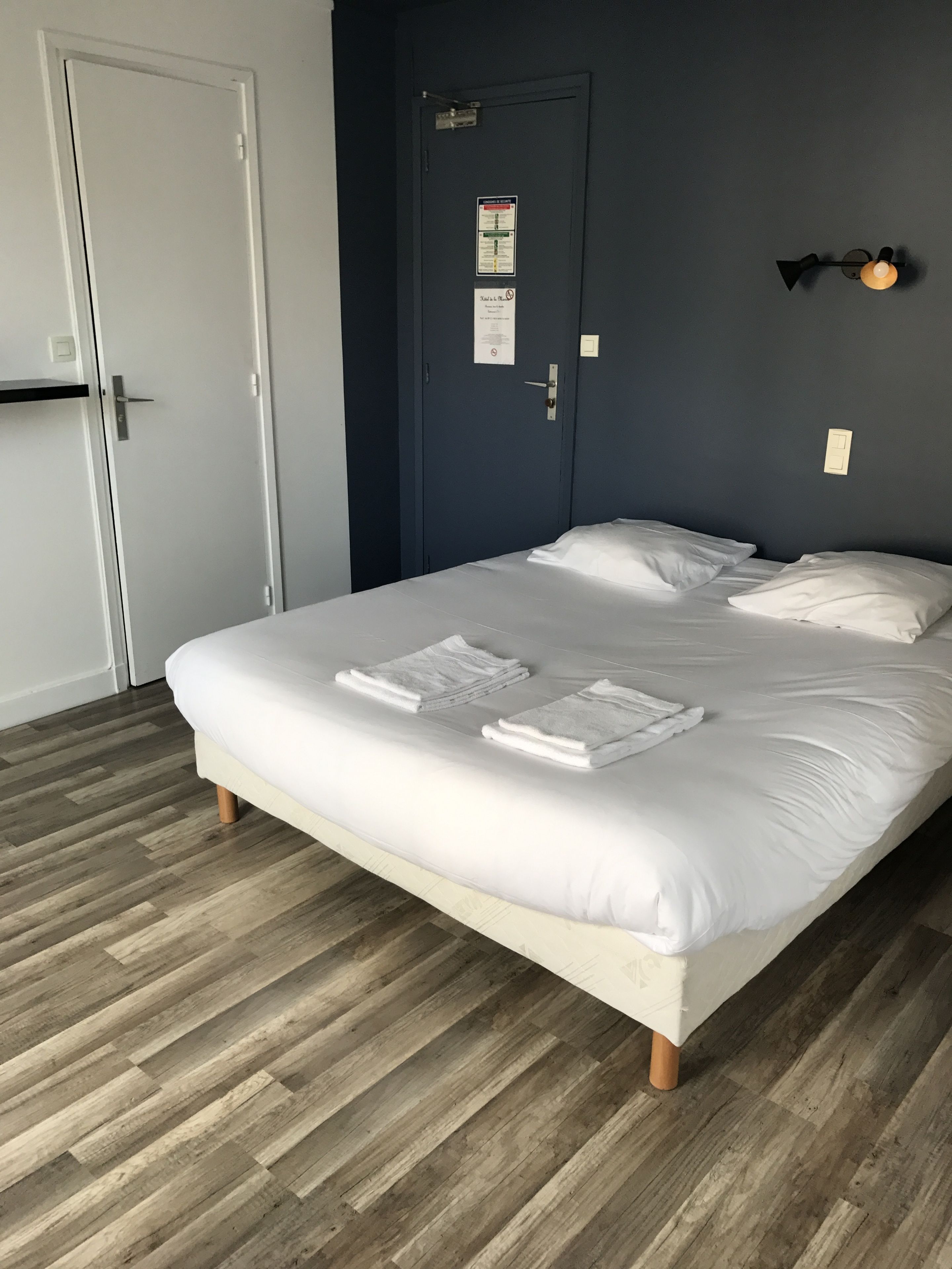 Una cama bien tendida con sábanas y toallas blancas, una pared azul oscuro, una puerta con un cartel y un interruptor de luz.