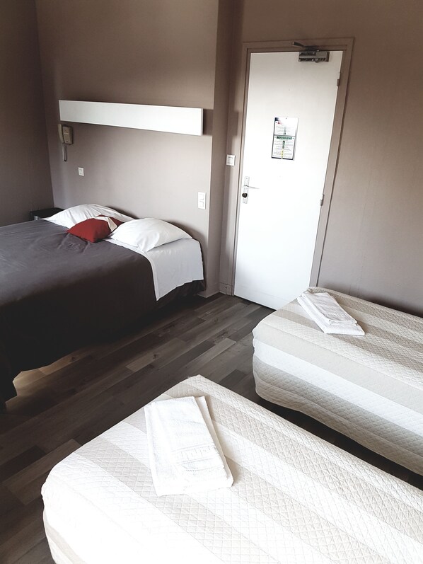 A room with two beds, one with a red pillow and the other with a white pillow. There is a door with a sign on it and a small table with a lamp. - Hôtel La Marine (Carnac)