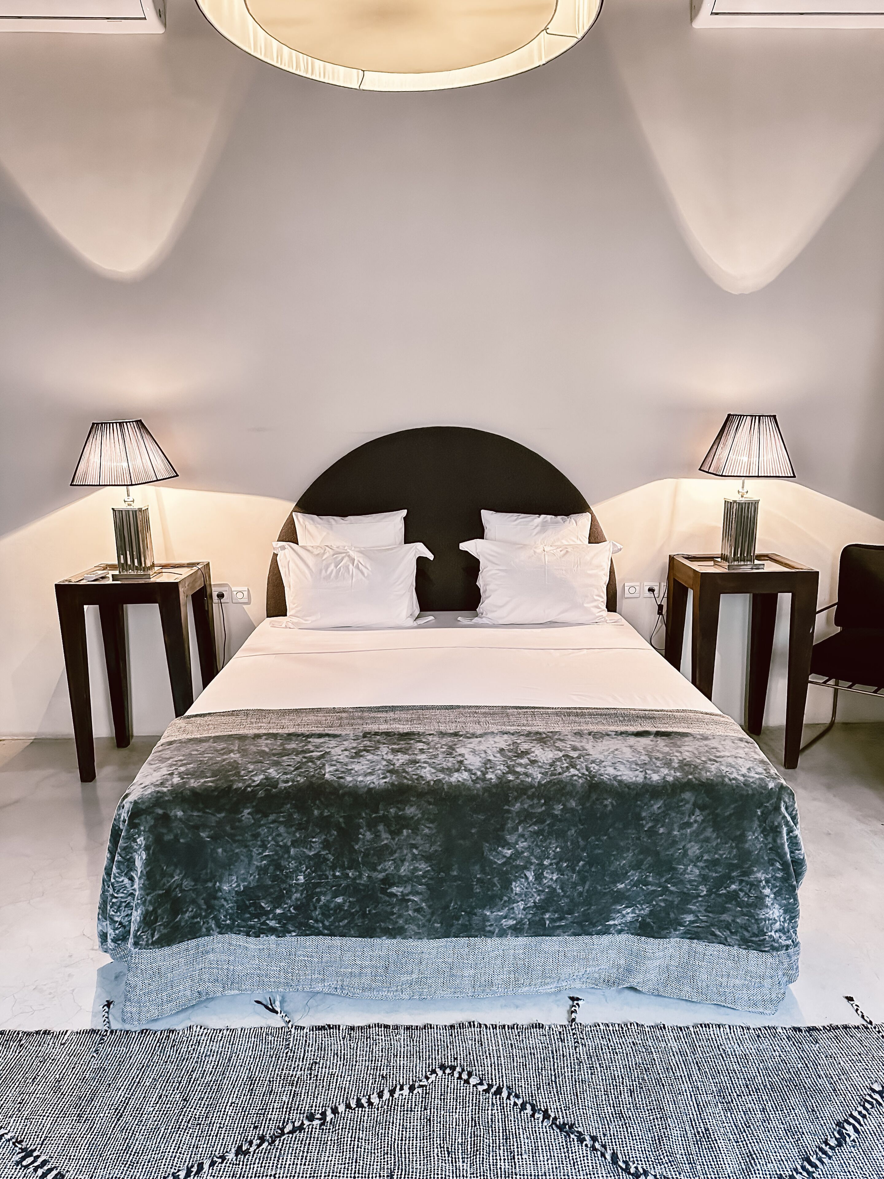 Une chambre d’hôtel moderne avec un grand lit, deux tables de chevet avec des lampes, une tête de lit sombre et un luminaire circulaire au-dessus du lit.