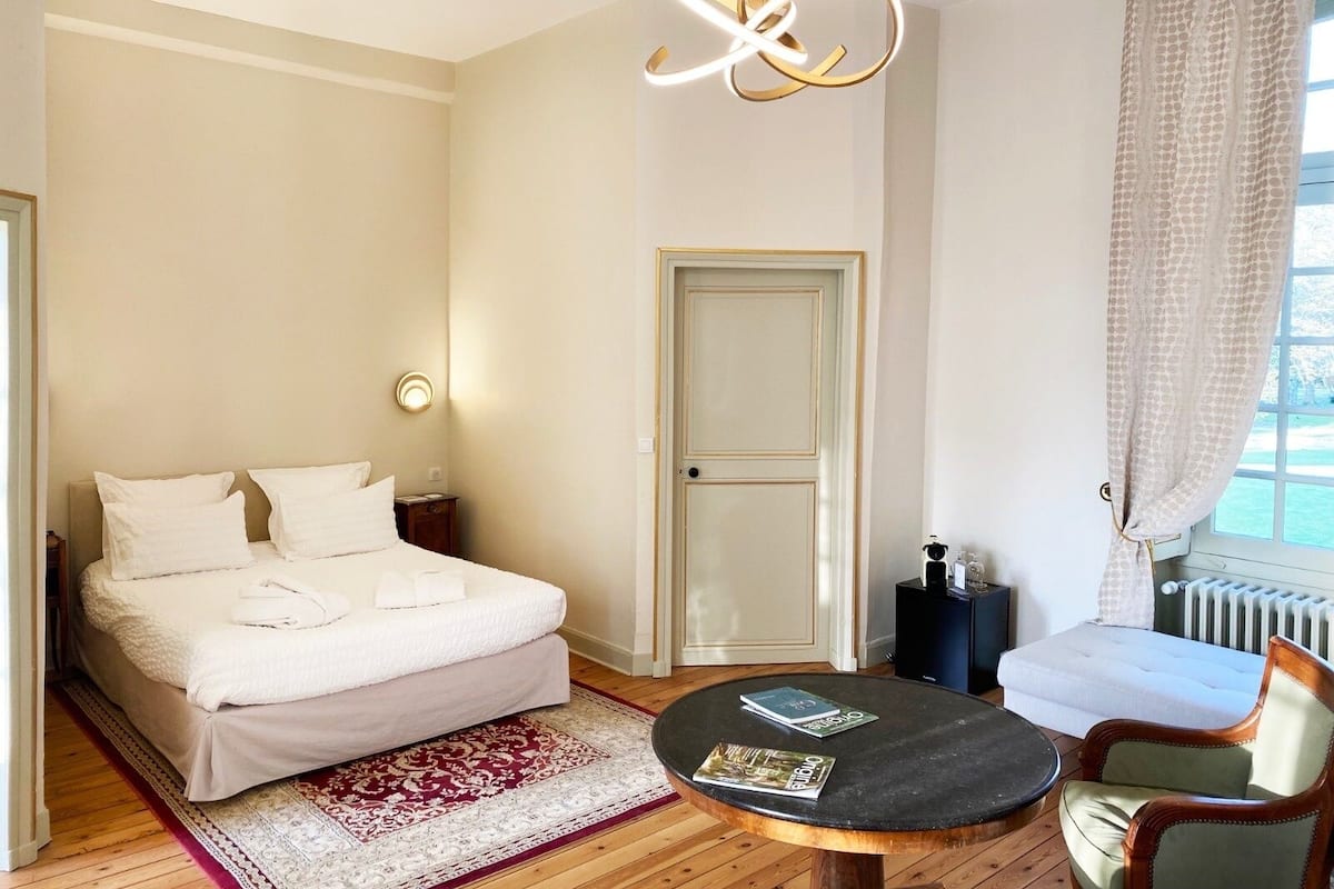 Una habitación de hotel con una cama, una mesita redonda con revistas, una silla, una mesita de noche, una lámpara y vistas al exterior a través de una ventana.