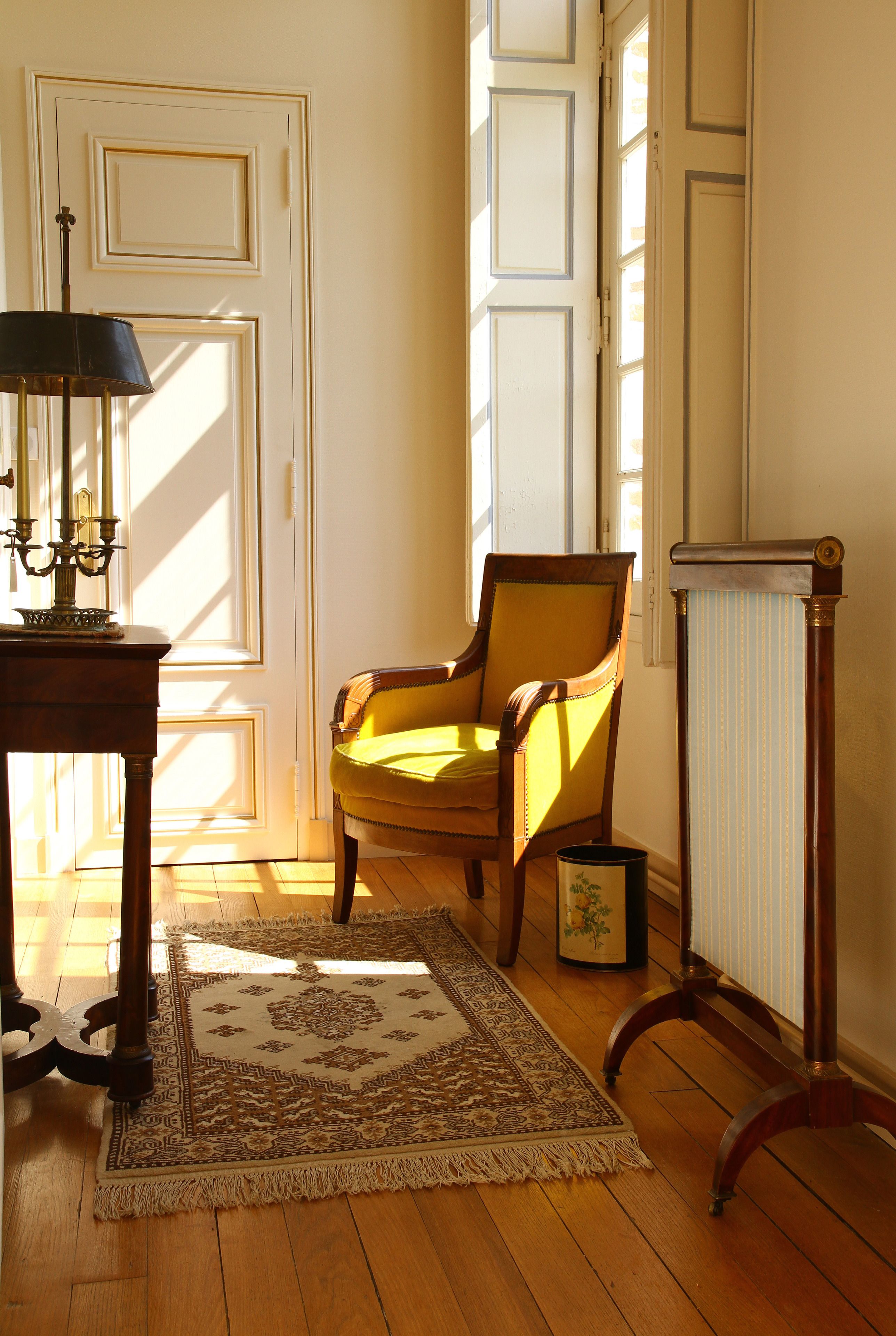 A room with a wooden floor, a yellow armchair, a patterned rug, a table with a lamp, and a radiator.
