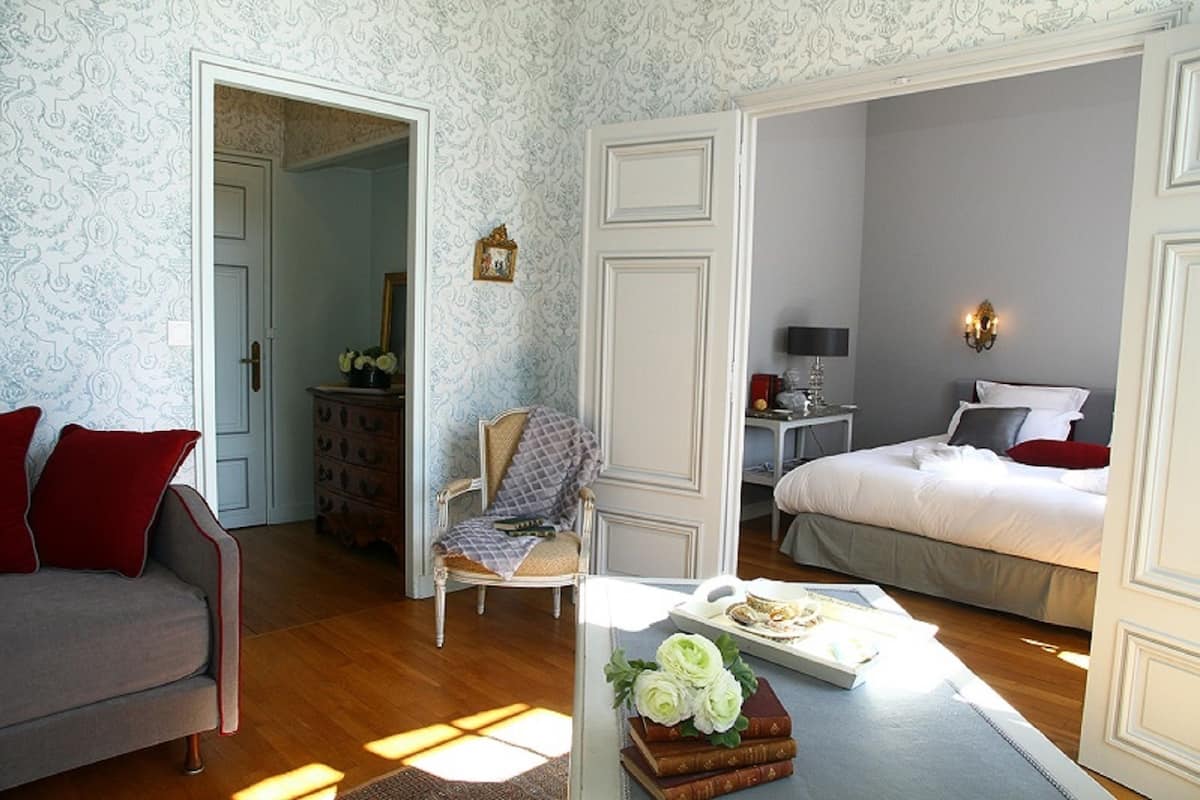 Un dormitorio con cama, una silla, una mesita con libros y flores, y un aparador.