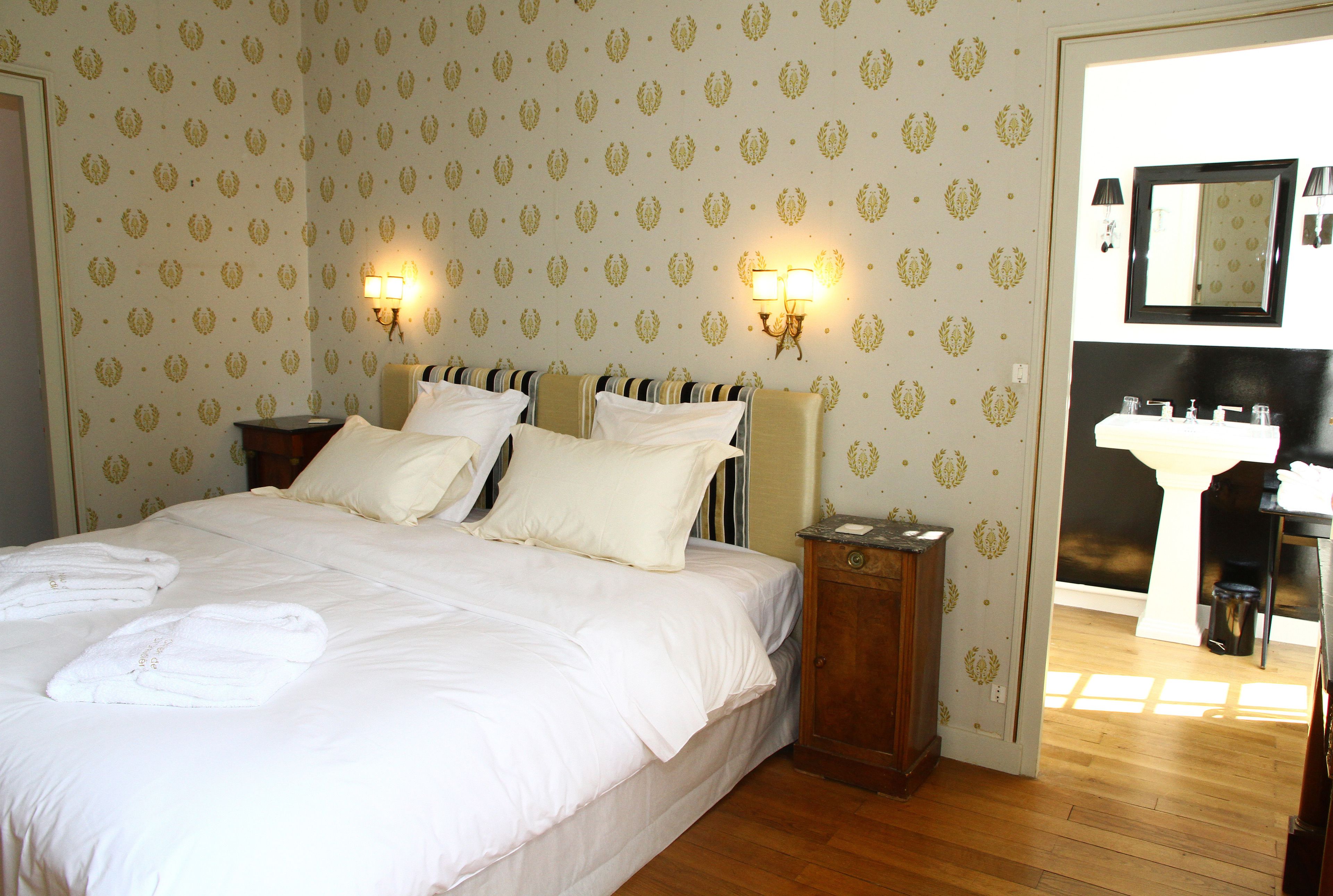 Ein Hotelzimmer mit einem großen Bett, zwei Nachttischlampen, einem Holznachttisch und einem Badezimmer mit Waschbecken und Spiegel.