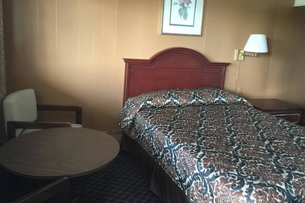 Quarto de hotel com cabeceira de madeira, mesa redonda, cama com colcha estampada, quadro pendurado na parede e abajur.