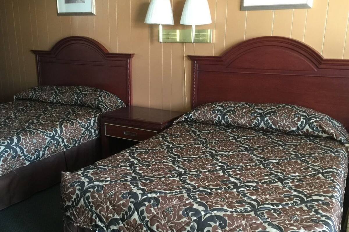 Quarto de hotel com duas camas, cada uma com roupa de cama estampada, cabeceira de madeira, mesa de cabeceira e luminárias fixadas na parede.