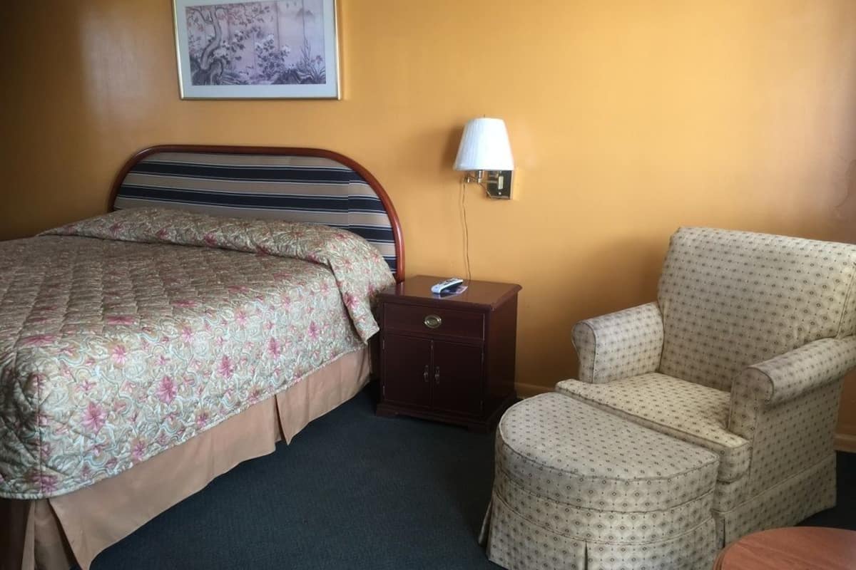 Quarto de hotel com cama, mesa de cabeceira, luminária, cadeira e pequena mesa.
