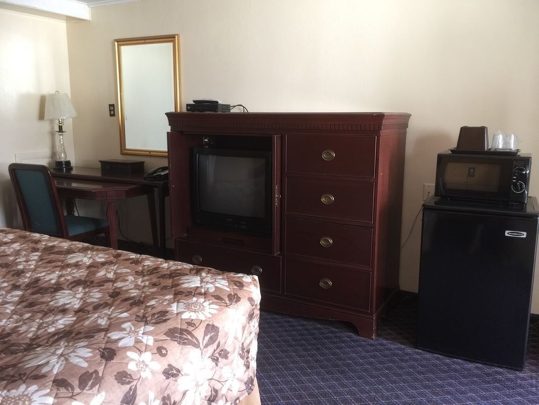 A hotel room with a bed, a desk with a lamp, a television, a dresser, and a refrigerator.