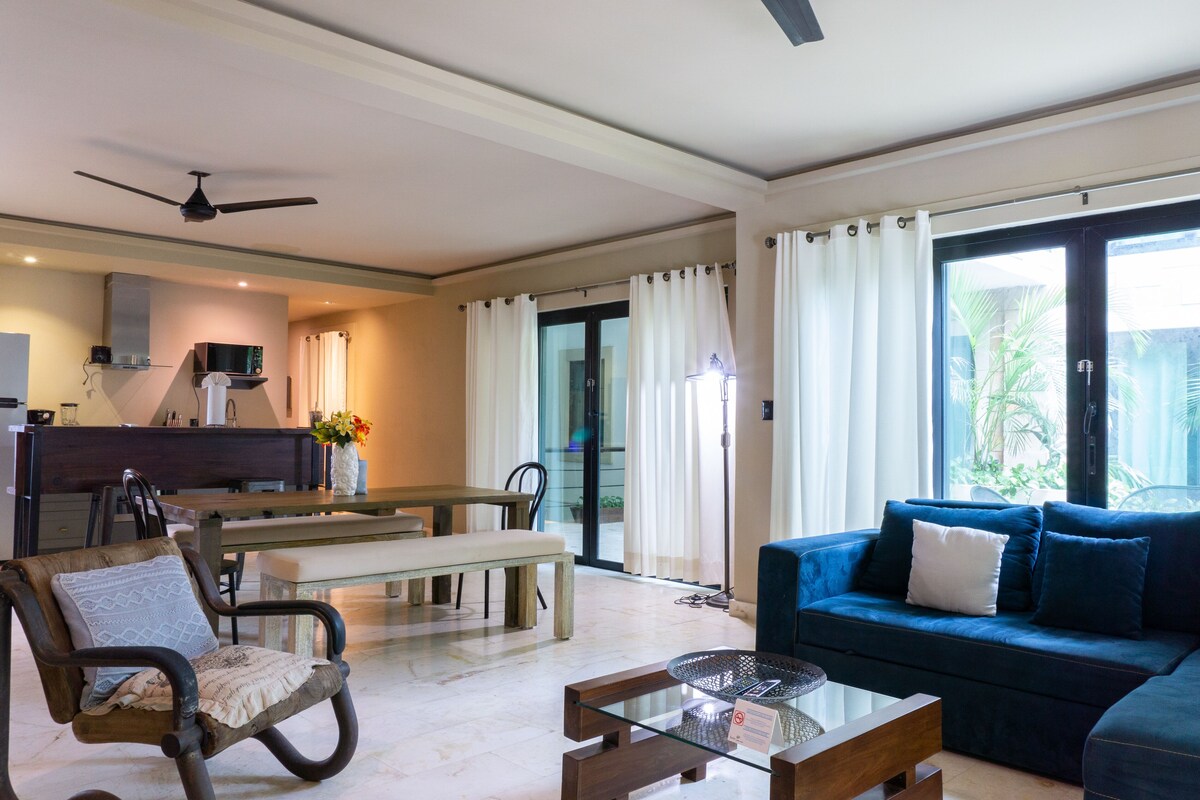 A living room with a blue sofa, a glass coffee table, a wooden bench, and a dining area with a table and chairs.