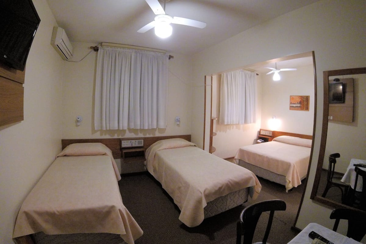 A hotel room with two single beds, a ceiling fan, an air conditioning unit, a bedside table with a lamp, and a mirror.
