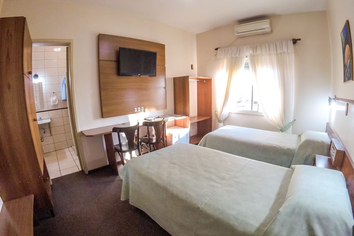 A hotel room with two beds, a desk with chairs, a TV mounted on the wall, an air conditioning unit, and a window with curtains.