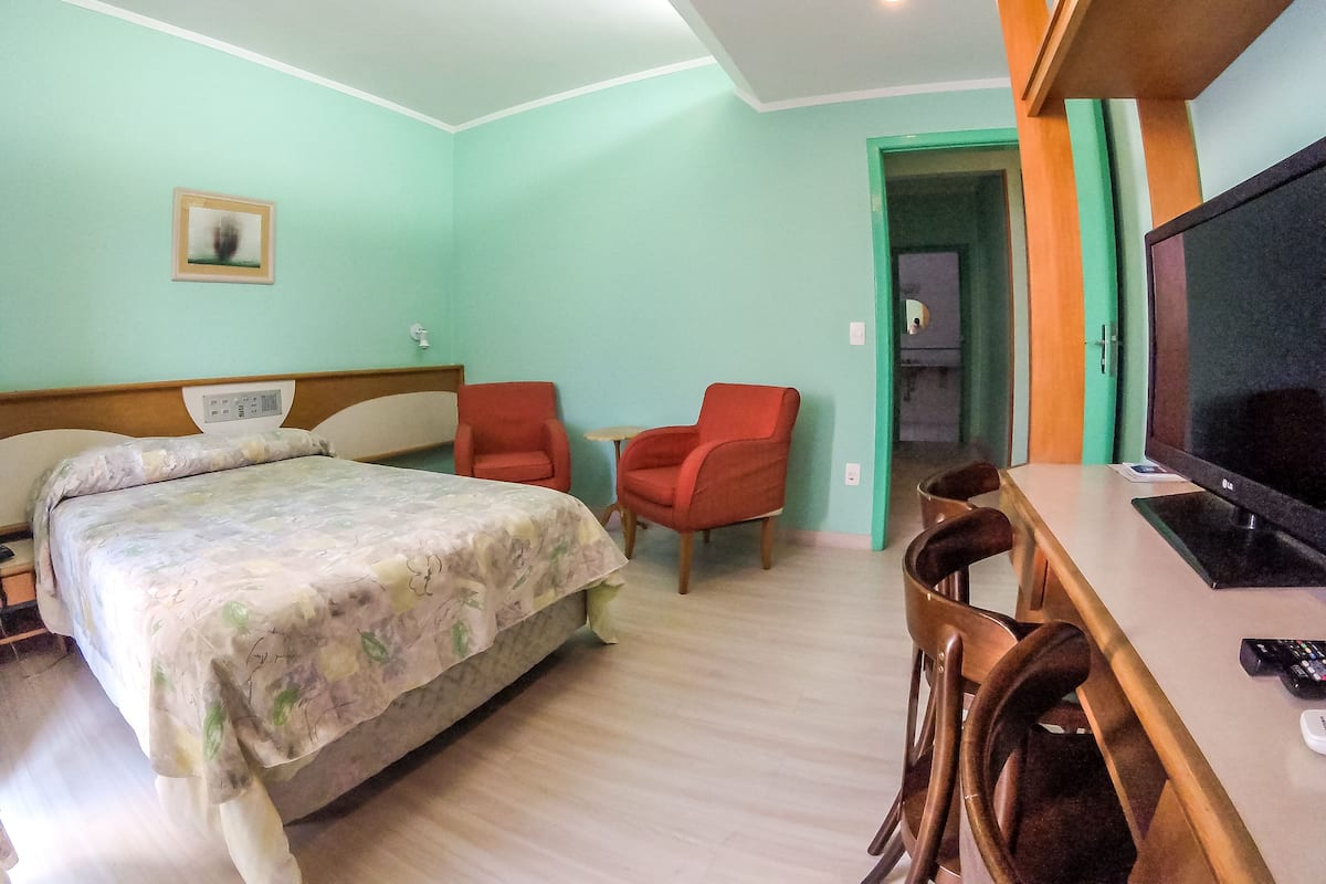 A hotel room with a bed, two red chairs, a desk with a television, and a door leading to another room.